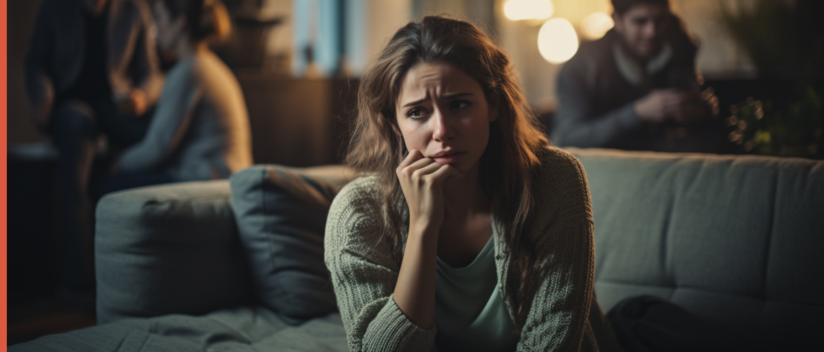 A woman looks anxious as she sits alone while friends socialize in the background.'