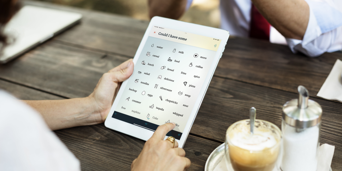 A woman's hand taps on a food-related word on an iPad running Spoken AAC. The woman is sitting at a dining table across from a man and there is a coffee and creamer to her right.