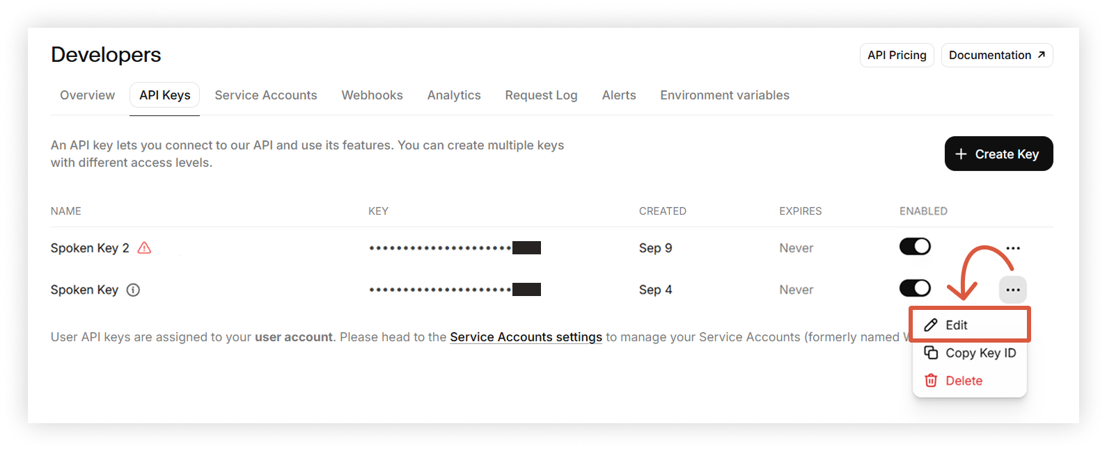 A screenshot of the Developers page on the ElevenLabs website, showing a list of API keys. An orange arrow points from the three dots mentioned in the tutorial to an open menu below with the options Edit, Copy Key ID, and Delete.